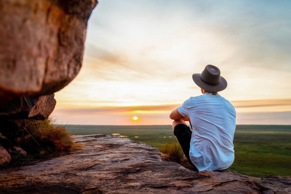 Sunset-Serenity-at-Ubirr-Kakadu A man stands on a rocky outcrop at Ubirr in Kakadu National Park, gazing at the breathtaking sunset. The vast, golden-hued sky stretches out above him, blending into fiery orange and soft pink tones that reflect across the wetlands below.