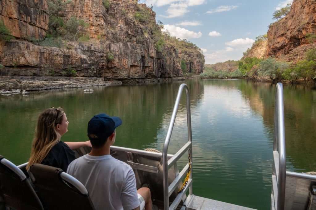 Nitmiluk-Gorge-Boat-Cruise-Katherine-National-Park A scenic boat cruise through Nitmiluk Gorge in Katherine National Park, showcasing towering sandstone cliffs and calm, reflective waters. Passengers on the boat enjoy breathtaking views of the ancient landscape, with lush greenery and the vibrant colors of the gorge adding to the serene and majestic experience.